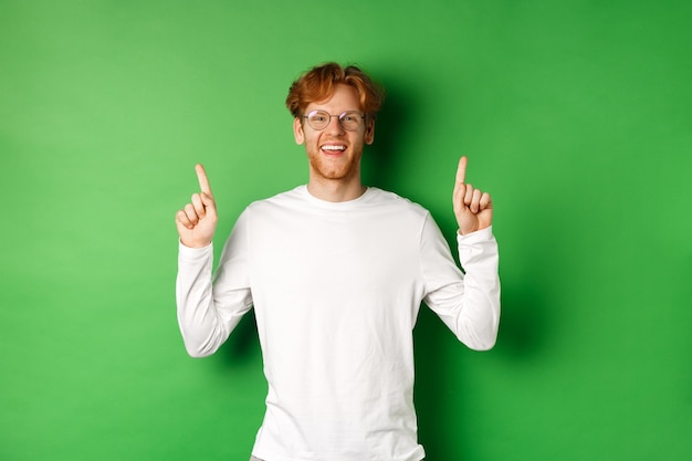 Foto glücklicher junger hipster mit roten haaren, brille tragend, finger nach oben zeigend und beförderung zeigend, in die kamera lächelnd, über grünem hintergrund stehend.