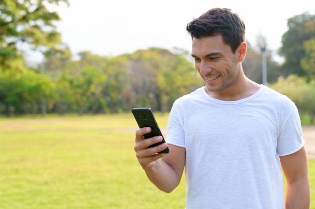 Glücklicher junger hübscher hispanischer Mann, der Telefon im Park im Freien verwendet