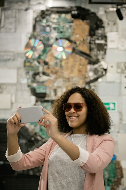 Glücklicher junger interkultureller weiblicher Tourist mit Smartphone, das Selfie auf der Außenseite des modernen Gebäudes macht