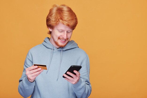 Glücklicher junger lässiger Mann mit Smartphone, der durch Waren im Online-Shop rollt und mit Kreditkarte bezahlt, während Bestellung macht