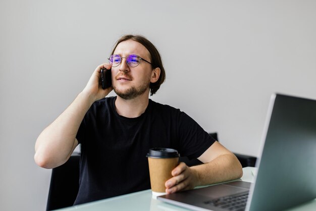 Glücklicher junger Mann, der am Handy spricht und Laptop im Büro benutzt