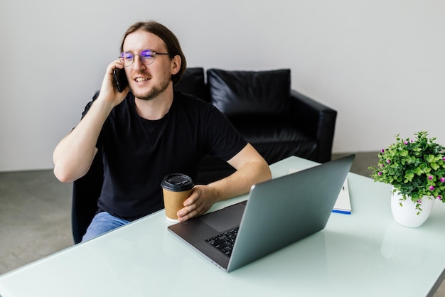 Glücklicher junger Mann, der am Handy spricht und Laptop im Büro benutzt