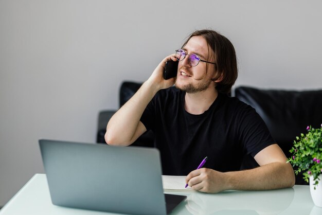 Glücklicher junger Mann, der am Handy spricht und Laptop im Büro benutzt