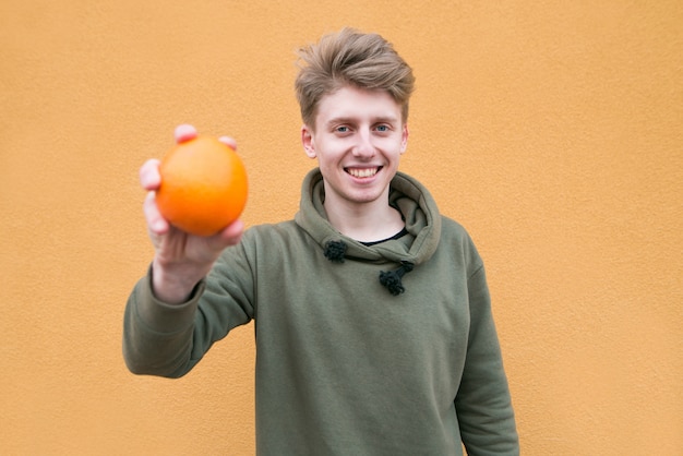 Glücklicher junger Mann, der auf einer orange Wand mit einer Orange in seinen Händen steht und lächelt. Konzentrieren Sie sich auf die Person