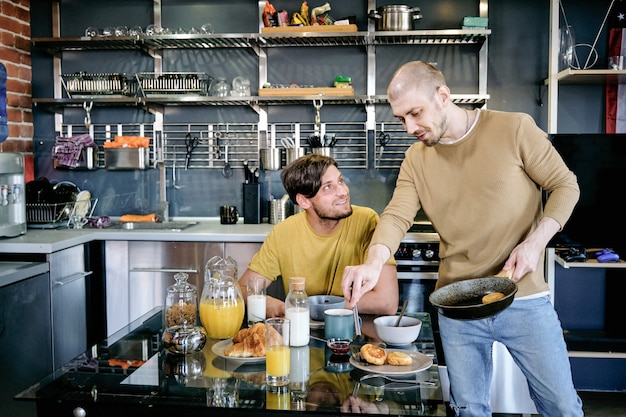 Glücklicher junger Mann, der beim Frühstück seinen Freund mit Bratpfanne anschaut