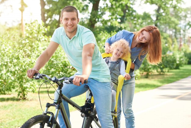 Glücklicher junger Mann, der beim Radfahren mit seinem Kind in einem Babyfahrradsitz und seiner schönen Frau am örtlichen Park auf einem aktiven Sommertagfamilien-aktiven Lebensstilkonzept lächelt.