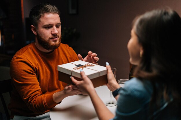 Glücklicher junger Mann, der ein Geschenk von einer liebevollen Frau erhält, die ein romantisches Abendessen mit Kerzen am Geburtstag oder am Valentinstag genießt