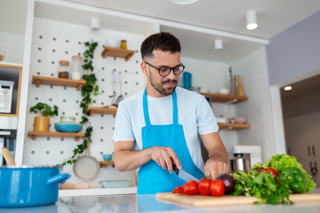 Glücklicher junger Mann, der frisches Gemüse oder Salatblätter hackt und vegetarischen Salat zubereitet, genießt das Kochen allein in der modernen Küche, die am Hobby-Aktivitätskonzept der Holzarbeitsplatte steht