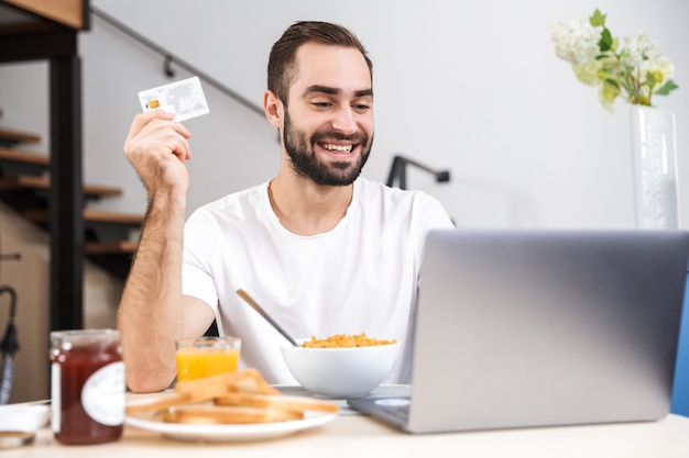 Glücklicher junger Mann, der Frühstück in der Küche, mit Laptop-Computer, zeigt Kreditkarte
