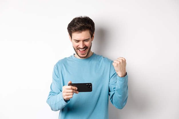 Glücklicher junger Mann, der im Videospiel auf dem Smartphone gewinnt, auf mobilen Bildschirm schaut und ja sagt, Faustpumpen in Feier macht, Onlineziel erreicht, über weißem Hintergrund stehend.