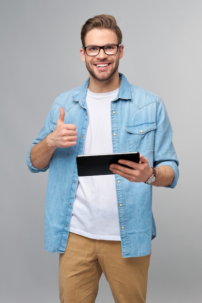 Glücklicher junger Mann, der Jeanshemd steht und Tablette über Studio graue Wand trägt