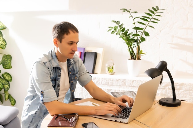 Glücklicher junger Mann, der lächelt, während er an seinem Laptop arbeitet, um alle seine Geschäfte frühmorgens mit seiner Tasse Kaffee zu erledigen