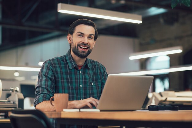 Glücklicher junger Mann, der mit einem Laptop bei der Arbeit sitzt und in die Ferne schaut