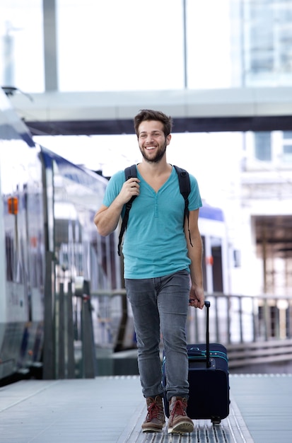 Glücklicher junger Mann, der mit Koffer an der Bahnstation geht