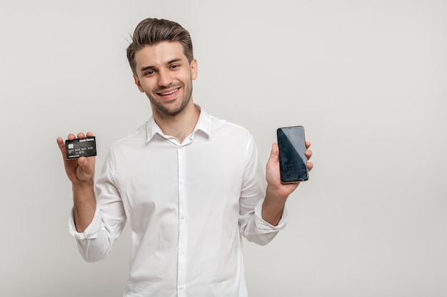 Foto glücklicher junger mann, der plastikkreditkarte zeigt, während er handy mit schwarzem bildschirm hält