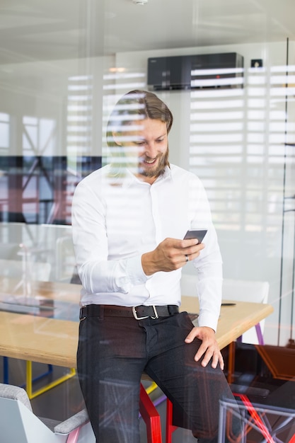 Glücklicher junger Mann, der sein intelligentes Telefon im Büro verwendet