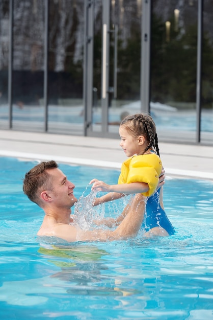 Glücklicher junger Mann, der seine süße kleine Tochter hebt, während er zusammen Spaß im Schwimmbad in einem modernen Sport- oder Freizeitzentrum hat