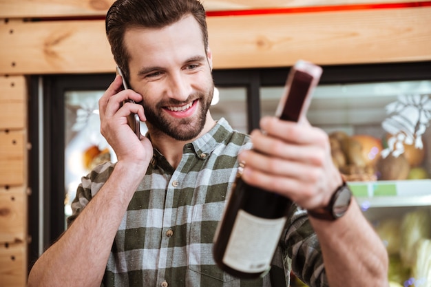 Glücklicher junger Mann, der Wein kauft und im Lebensmittelgeschäft mit dem Handy spricht