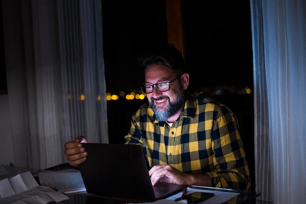 Glücklicher junger Mann, der zu Hause einen Laptop oder Computer verwendet und spät in der Nacht im Dunkeln arbeitet, mit dem Licht des Bildschirms auf seinem Gesicht, lächelnd xA