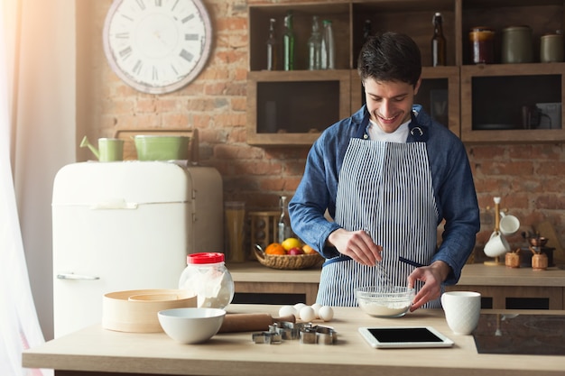 Glücklicher junger Mann, der zu Hause Kuchen in der Dachbodenküche backt, mit digitalem Tablet mit Rezept. Mockup, Kopienraum