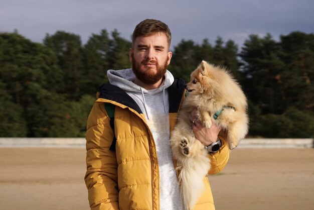 Glücklicher junger Mann geht mit seinem Haustier Pomeranian Spitz-Hund am Strand und hält einen Welpen in den Händen