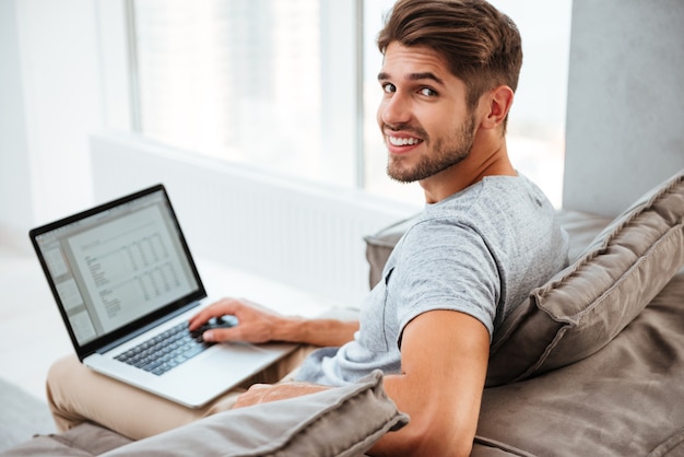 Glücklicher junger Mann im T-Shirt, der auf Sofa zu Hause sitzt. Arbeiten am Laptop und Lächeln beim Betrachten der Kamera.