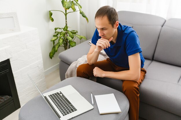 Glücklicher junger Mann im T-Shirt, der zu Hause sitzt, an Laptop-Computer arbeitet, lächelnd.