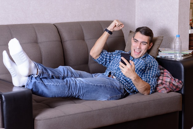 Glücklicher junger Mann in den Jeans, in einem karierten Hemd und in weißen Socken, die auf der Couch mit einem Smartphone liegen und ist glücklich, seine Hand oben anzuheben