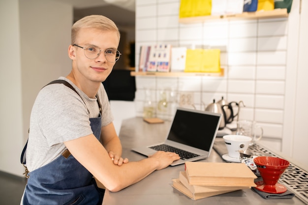 Glücklicher junger Mann in der Uniform und in den Brillen, die am Arbeitsplatz stehen und Sie beim Lernen vor Laptop betrachten