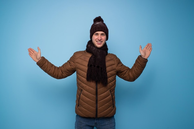 Foto glücklicher junger mann in einem braunen schal und einer wintermütze, eine hellbraune jacke auf blauem hintergrund breitete seine arme zu den seiten aus