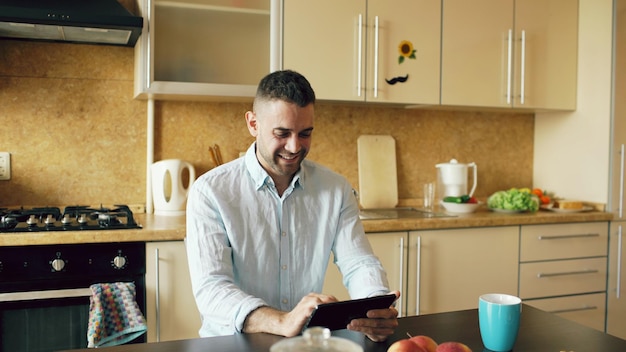 Glücklicher junger Mann mit digitalem Tablet-Computer, der zu Hause in der Küche sitzt