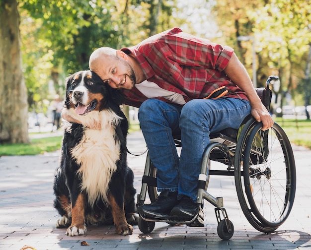 Glücklicher junger Mann mit einer körperlichen Behinderung, der mit seinem Hund einen Rollstuhl benutzt