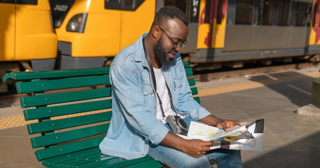 Glücklicher junger Mann mit Karte, der am Bahnhof sitzt