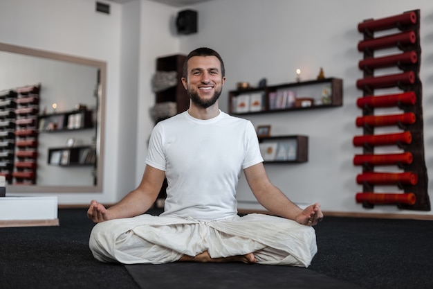 Glücklicher junger Mann mit positivem Lächeln mit Bart, der Yoga im Fitness-Studio praktiziert. Gesunder Lebensstil.