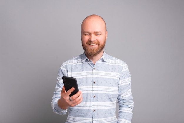 Glücklicher junger Mann mit Smartphone, der in die Kamera schaut und über grauem Hintergrund lächelt