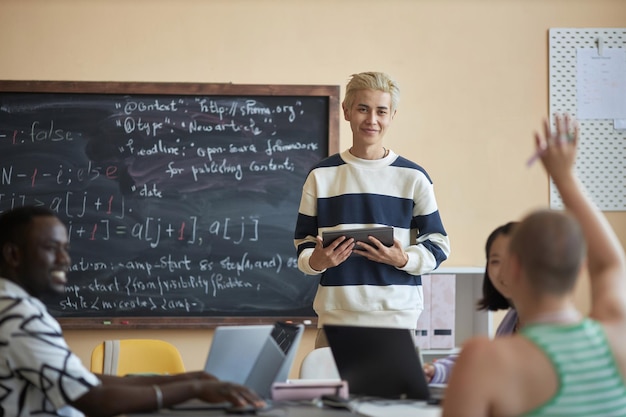 Glücklicher junger Mann mit Tablet, der eine Studentin ansieht, die während der Präsentation im Informatikunterricht die Hand hebt, um eine Frage zu stellen