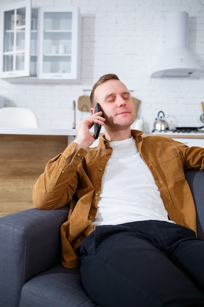 Glücklicher junger Mann sitzt mit einer Tasse Kaffee auf dem Sofa im Wohnzimmer und telefoniert.