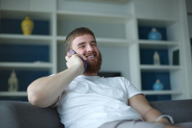 Glücklicher junger Mann spricht Chat auf Mobiltelefon Lächeln Spaß haben sitzen auf der bequemen Couch zu Hause in