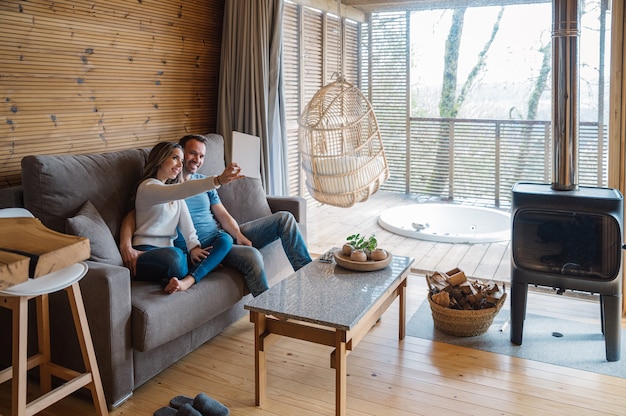 Glücklicher junger Mann und Frau in der Freizeitkleidung, die auf Sofa sitzt und selfie auf Tablette nimmt, während Freizeit zusammen im gemütlichen Wohnzimmer im rustikalen Stil verbringen