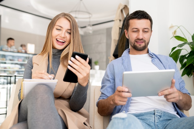Glücklicher junger Student, der Smartphonebildschirm betrachtet, während Notizen im Notizblock mit ihrem Freund macht, der Touchpad in der Nähe verwendet