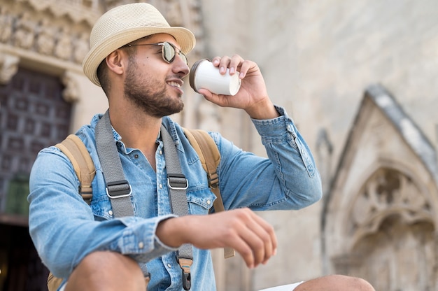 Glücklicher junger Touristenmann mit Kaffee zum Mitnehmen und Rucksack, der auf der Straße ruht?