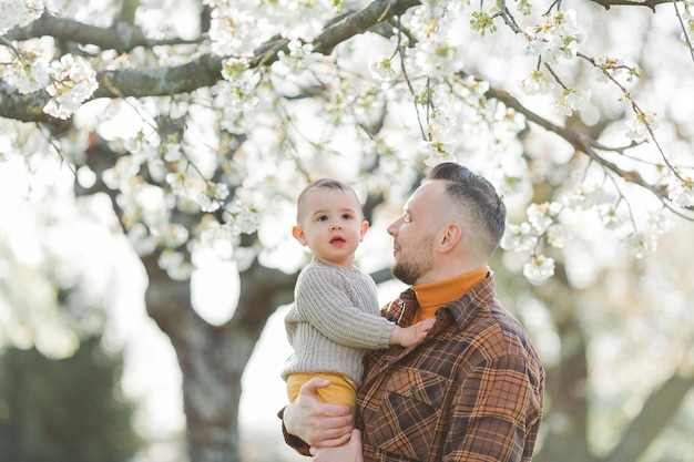 Glücklicher junger Vater mit kleinem Sohn spaziert durch blühenden Garten Vater mit 1-jährigem Baby Frühling