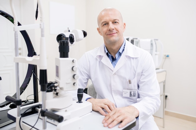 Glücklicher kahler Augenarzt mittleren Alters im weißen Kittel, der am Arbeitsplatz durch medizinische Ausrüstung in seinen Kliniken sitzt