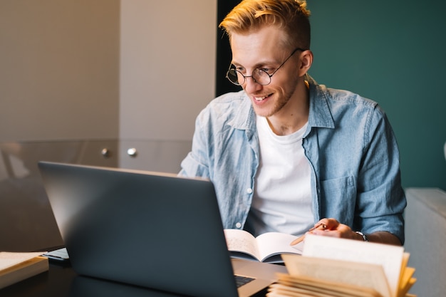 Glücklicher kaukasischer Mann-College-Student in den Gläsern, die mit Laptop studieren, der entfernt entfernt für Testprüfungs-Aufsatz arbeitet, der Hausaufgaben zu Hause macht