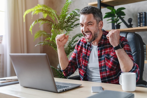 Glücklicher kaukasischer Mann, der im Heimbüro sitzt, macht Siegergeste