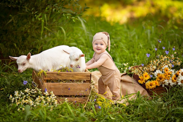 Glücklicher Kinderjunge mit weißen Ziegen im Frühjahr auf Natur im Dorf mit Grün und Blumen.