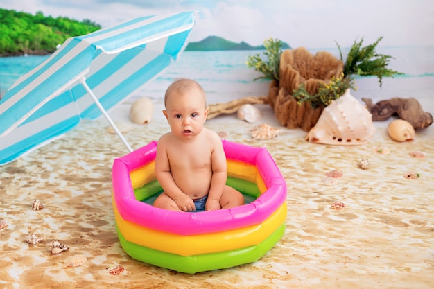 Glücklicher kleiner Junge badet in einem hellen aufblasbaren Pool an einem Sandstrand mit Palmen am Meer