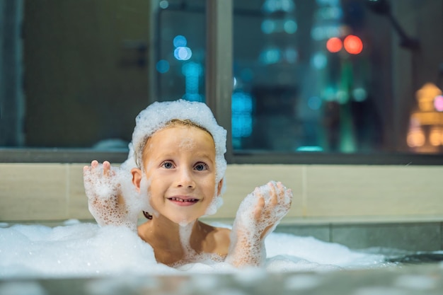 Glücklicher kleiner Junge, der abends in der Badewanne sitzt, bevor er auf dem Hintergrund eines Fensters mit Blick auf die Abendstadt schlafen geht. Porträt eines Babys, das in einem Bad voller Schaum in der Nähe des Fensters badet.