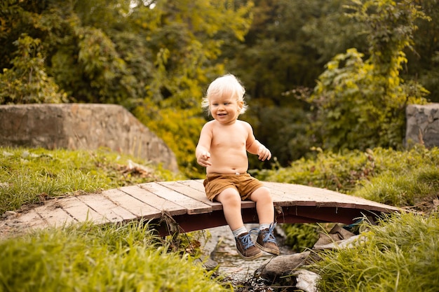 Glücklicher kleiner Junge, der auf der Brücke über dem Bach sitzt und im Herbst- oder Sommerpark mit goldenen Bäumen lächelt