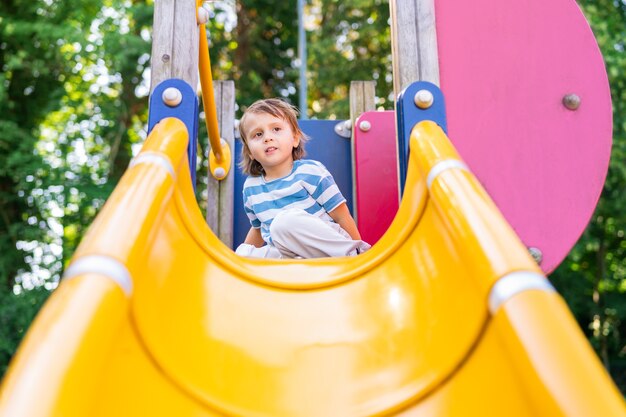 Glücklicher kleiner Junge, der in einem Park auf Spielplatz spielt. Kleinkind, das wegschaut, sitzt auf einer Rutsche. Sommeraktivitäten für Kinder.